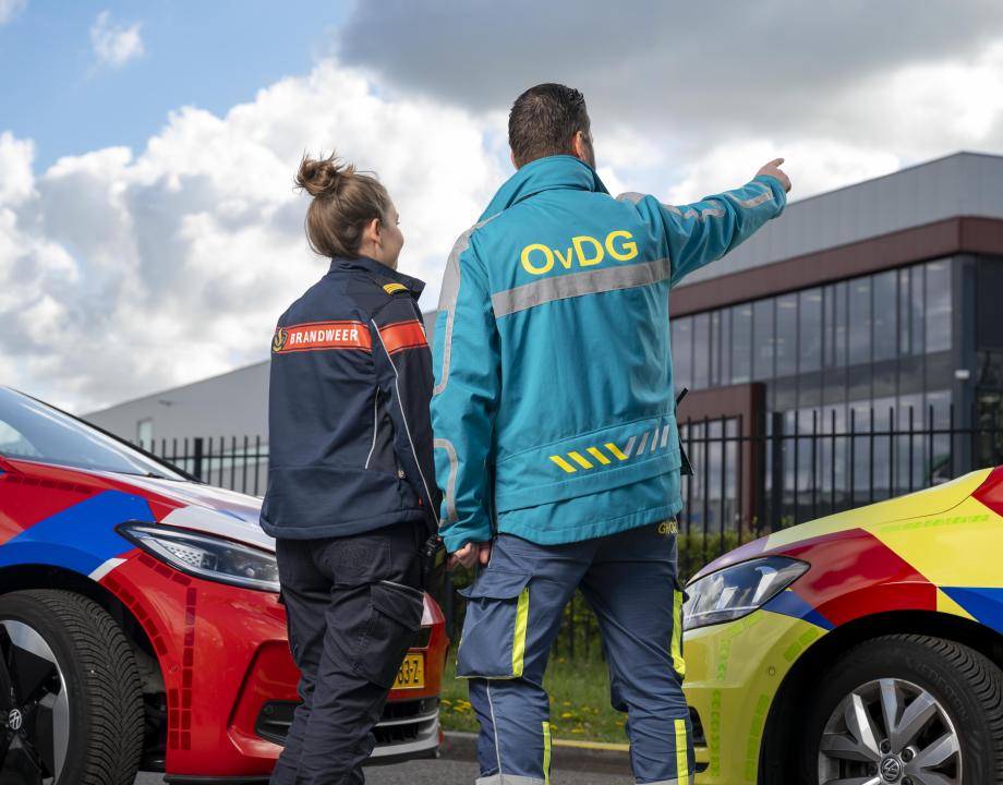 Brandweervrouw en GHOR medewerker staan tussen een ambulance en brandweerauto te kijken naar een gebouw